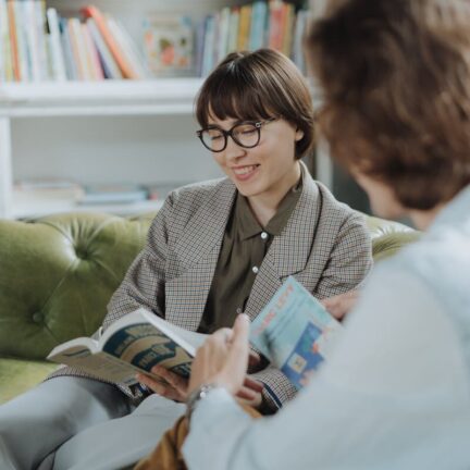Two young adults enjoy reading on a sofa in a cozy library setting, surrounded by books.